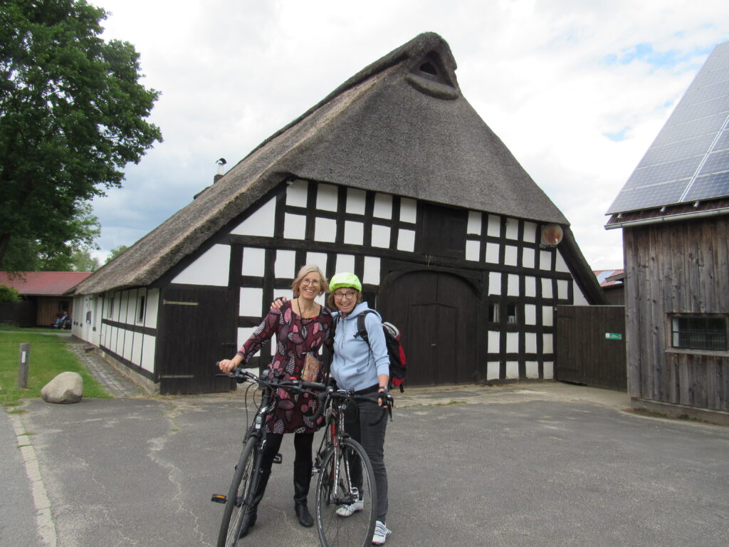 two women with bicycles standing in front of old house with thatched roof.