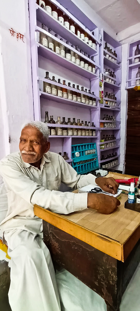 Man sitting in from of shop filled with rows of medicine bottles